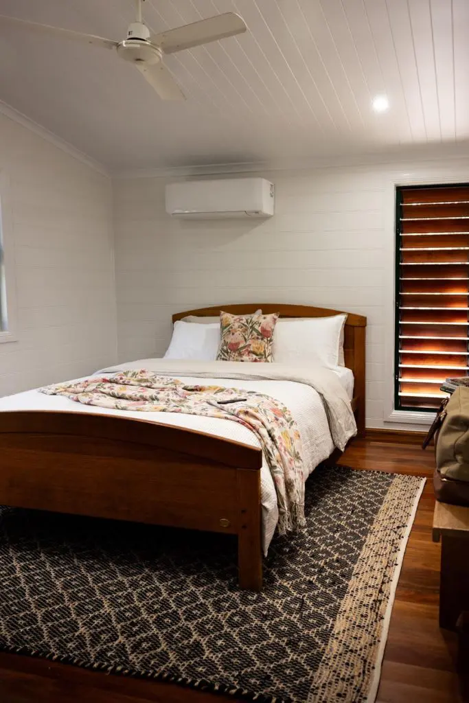 Master bedroom interior featuring a dark wood bed frame, white bedding with a floral runner, a black-and-white patterned rug, ceiling fan, and air conditioner inside Palmerston Cottage.