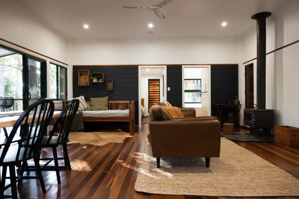 Open-plan living room interior at Palmerston Cottage featuring hardwood floors, a brown leather couch, a traditional wood-burning fireplace heater, and a dining table with chairs.
