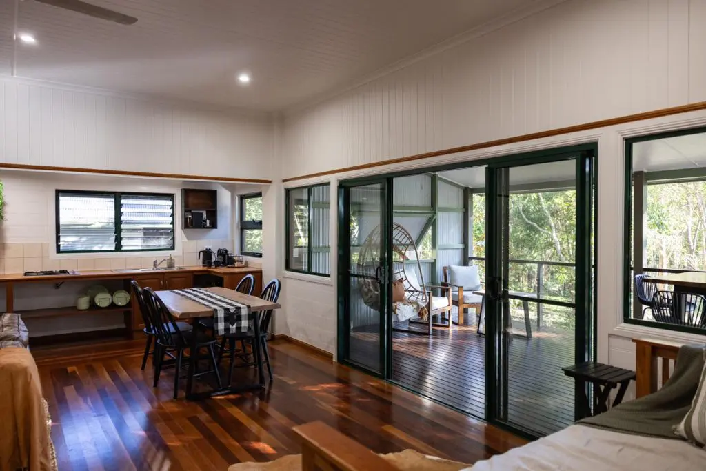 Interior view of Palmerston Cottage showing an open-plan kitchen and dining area opening out onto a timber deck with a hanging egg chair and rainforest view.
