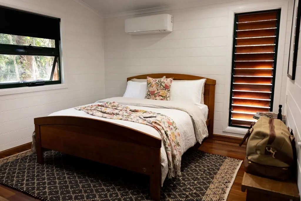 Master bedroom interior at Palmerston Cottage showing a wooden queen bed with floral bedding, air conditioner, shuttered window, and a leather travel bag on a bench.